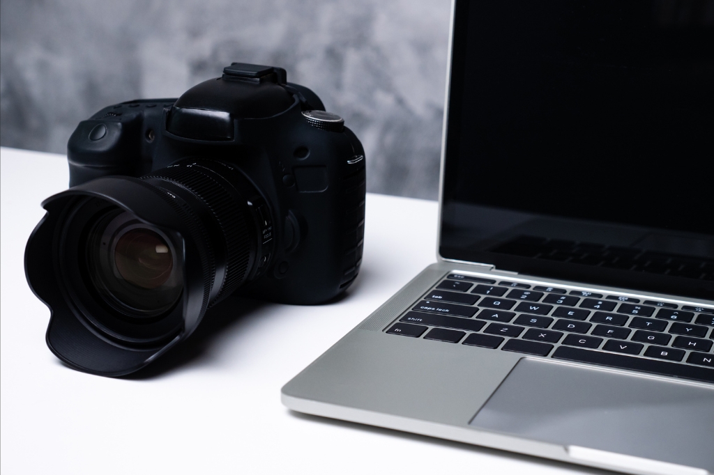 Close-up of a DSLR camera next to an open laptop on a white desk.