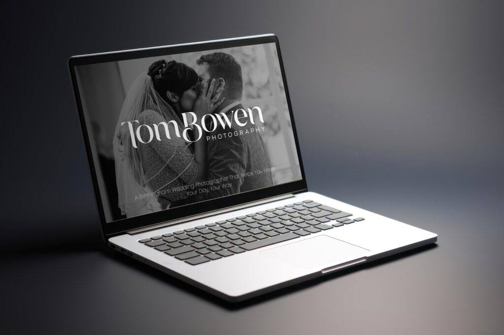 Laptop displaying a wedding photography website with the headline 'Tom Bowen Photography' and a black-and-white image of a bride and groom kissing. The tagline reads 'A Birmingham Wedding Photographer That Helps You Have Your Day, Your Way.'