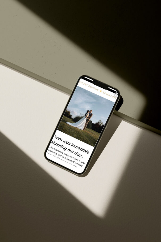 Mobile phone displaying a wedding photography website's review section, showing a photo of a bride and groom and a testimonial that reads, 'Tom was incredible shooting our day.' The phone is angled against a beige and shadowed background.