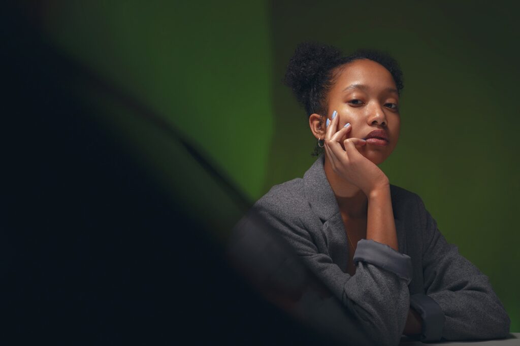 A young woman with natural hair styled in a bun sits at a table, resting her chin on her hand with a contemplative expression. She is wearing a grey blazer, and the background has a greenish hue that softly lights the scene.