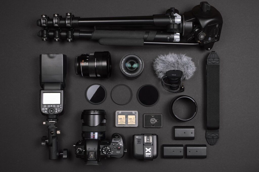 A flat lay of various camera equipment and accessories arranged neatly on a dark surface. Items include a tripod, camera lenses, a camera body, flash, microphone with a windscreen, memory cards, filters, batteries, and a camera strap. Highlighting the technical elements to photography and SEO