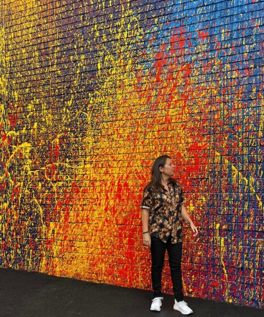 A person standing in front of a colourful graffiti wall, wearing a patterned shirt and white sneakers, looking off to the side.