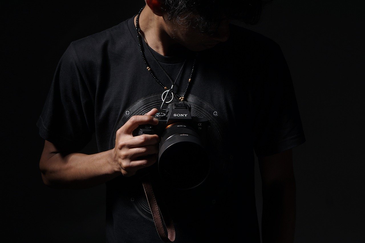 A photographer wearing a black t-shirt, holding a Sony camera, with their head slightly bowed and a focus on the camera in low light.