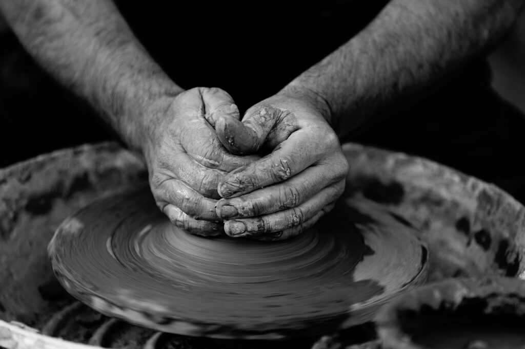 A close-up shot of a potter's hands working with wet clay on a spinning pottery wheel. The image is in black and white, emphasising the rough texture of the clay and the concentration in the potter’s hands as they shape the material.