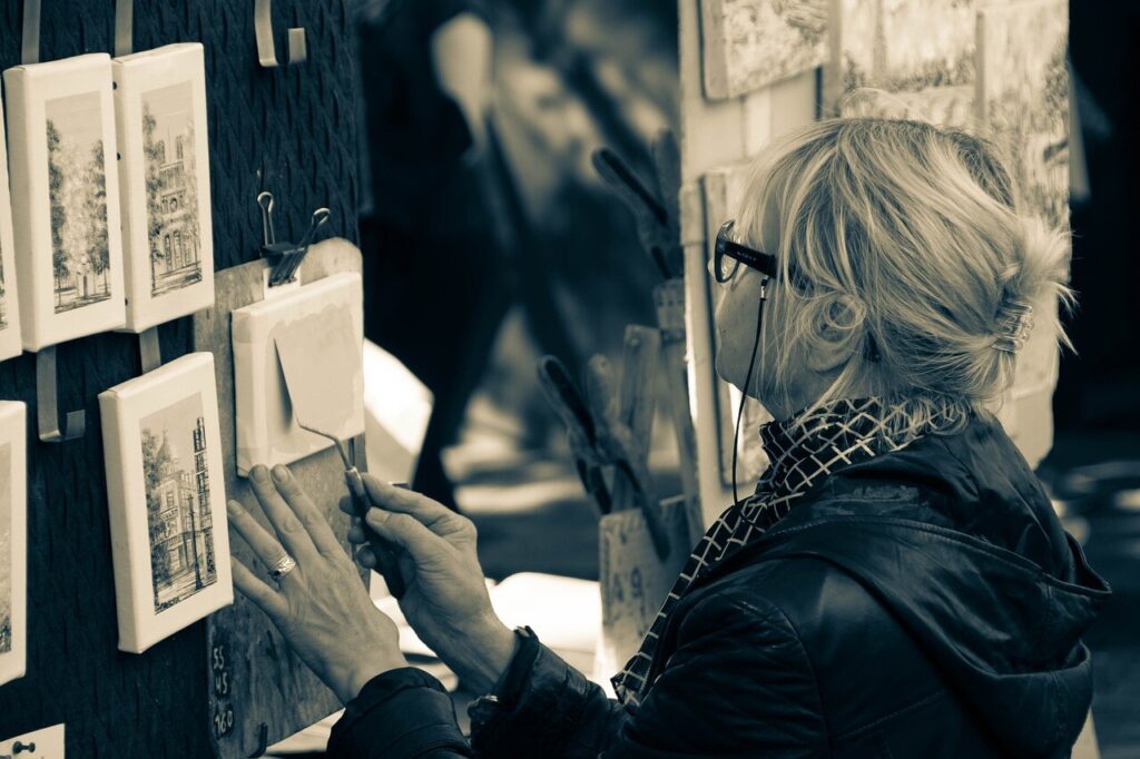 A woman with blonde hair, tied up in a clip, is seen from the side painting on small canvases attached to a display board. She is wearing glasses and a patterned scarf, focused on her work using a palette knife amidst a setting of other detailed paintings.