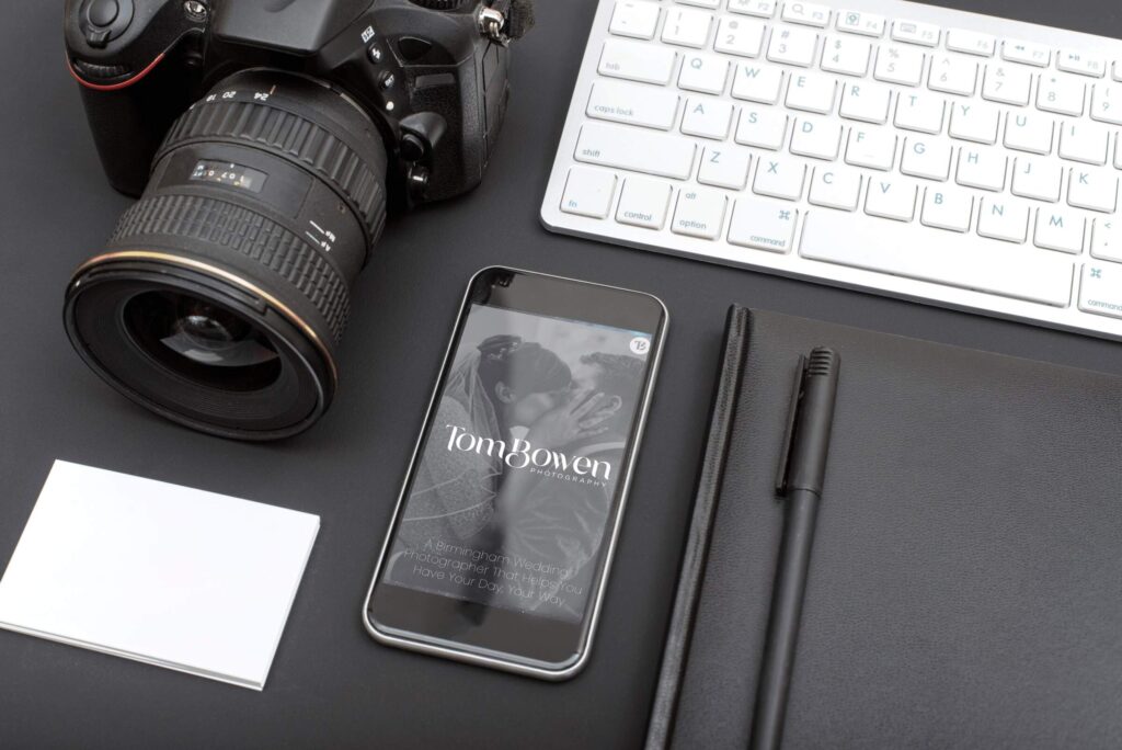 A flat lay of a photographer's workspace featuring a camera, a smartphone displaying a wedding photography website, a keyboard, a notebook, a pen, and business cards on a dark surface.