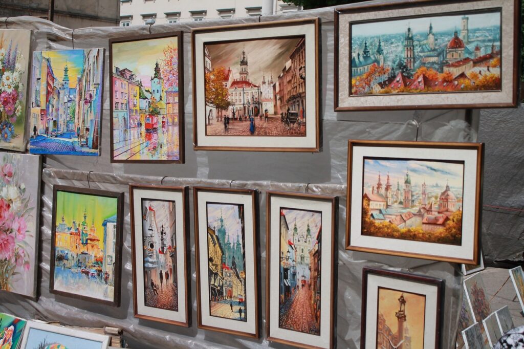 A display of vibrant, framed paintings at an outdoor exhibition. The artwork showcases cityscapes and landscapes in various styles, with buildings and colourful streets featured prominently.