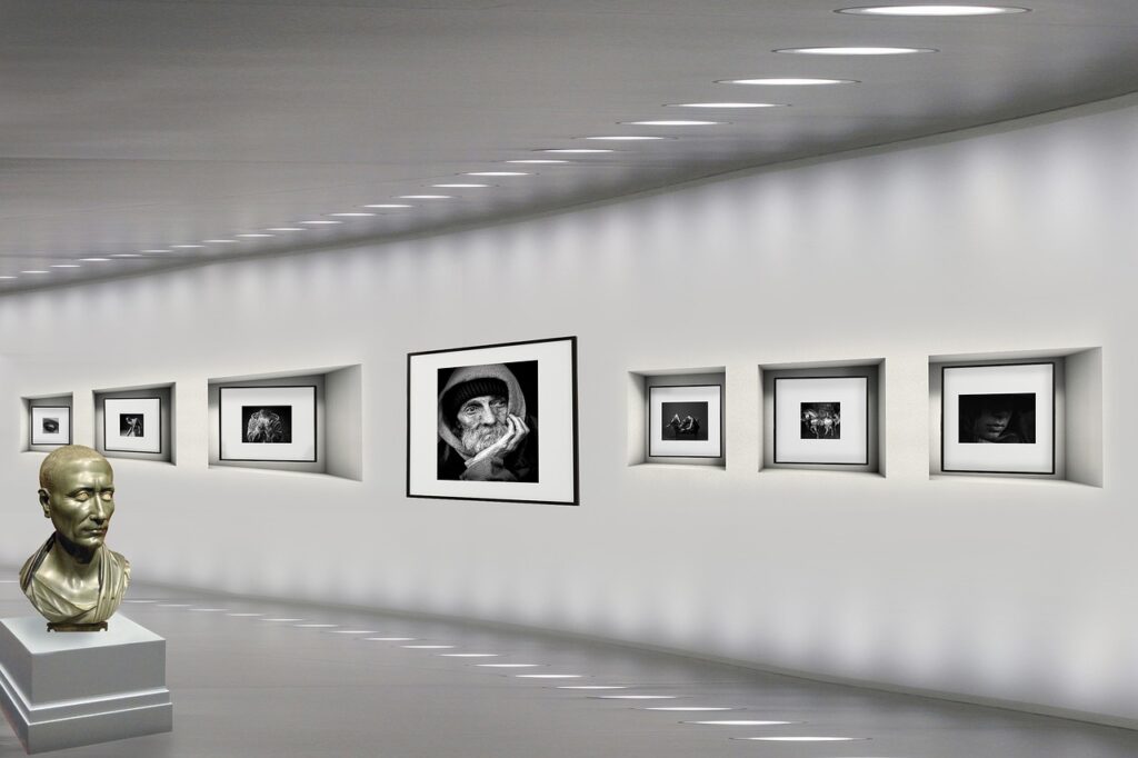 A sleek, modern art gallery with black-and-white photographs displayed along a minimalistic white wall. A bronze bust statue is placed on a pedestal at the entrance of the exhibit.