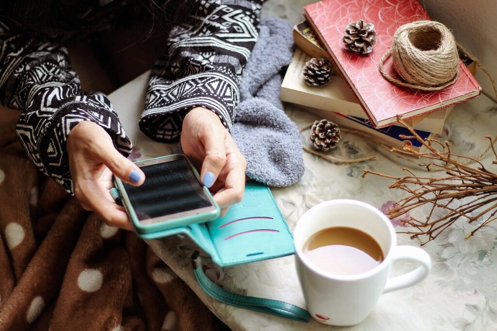 Close-up of an artist wearing a patterned sweater, using a smartphone with a turquoise case while seated at a cozy table. The table has a cup of coffee, pinecones, books, and craft supplies, creating a relaxed workspace atmosphere.