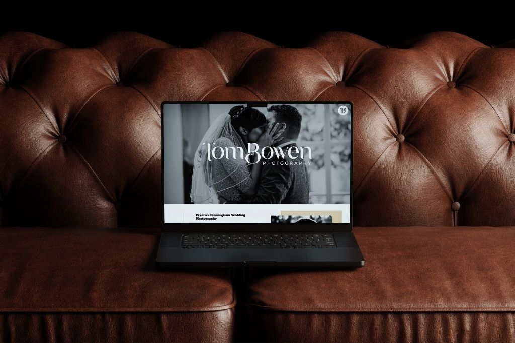 Laptop displaying Tom Bowen Photography website with a black-and-white wedding photo, placed on a brown leather tufted sofa.